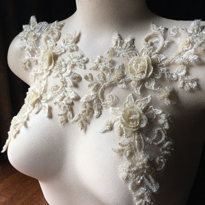 May include: A delicate ivory lace applique with floral designs and pearl-like embellishments. The applique features intricate floral patterns and delicate beading, perfect for adding a touch of elegance to a garment.