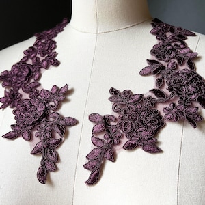 May include: Two plum-colored floral lace appliques. Each applique features detailed embroidered flowers and leaves, with a delicate net backing. The appliques are displayed on a mannequin, showcasing their potential use in garment design.