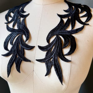 Könnte beinhalten: Ein Paar schwarze, bestickte Applikationen mit einem symmetrischen, länglichen Blatt- oder Flammenmuster. Diese dekorativen Stoffteile sind mit aufwendigen Stickereien sowie geschwungenen, spitzen Kanten versehen und liegen nebeneinander auf einer Form.