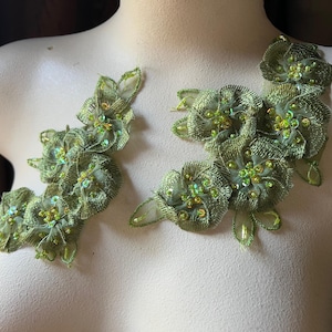 Puede incluir: Dos apliques florales verdes con detalles intrincados. Cada aplique presenta pétalos de tela en capas, adornados con pequeñas cuentas y lentejuelas en tonos verdes y dorados. Los apliques están diseñados para coser sobre tela.