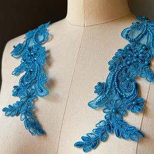 May include: Two turquoise lace appliques with floral and beaded designs. The appliques are displayed on a mannequin, showcasing their intricate details and potential for embellishment on garments or crafts.