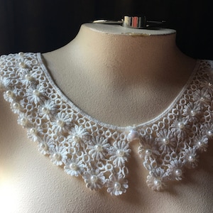 Puede incluir: Collar de encaje blanco con adornos de perlas. El collar est&aacute; en un busto de maniqu&iacute; blanco.