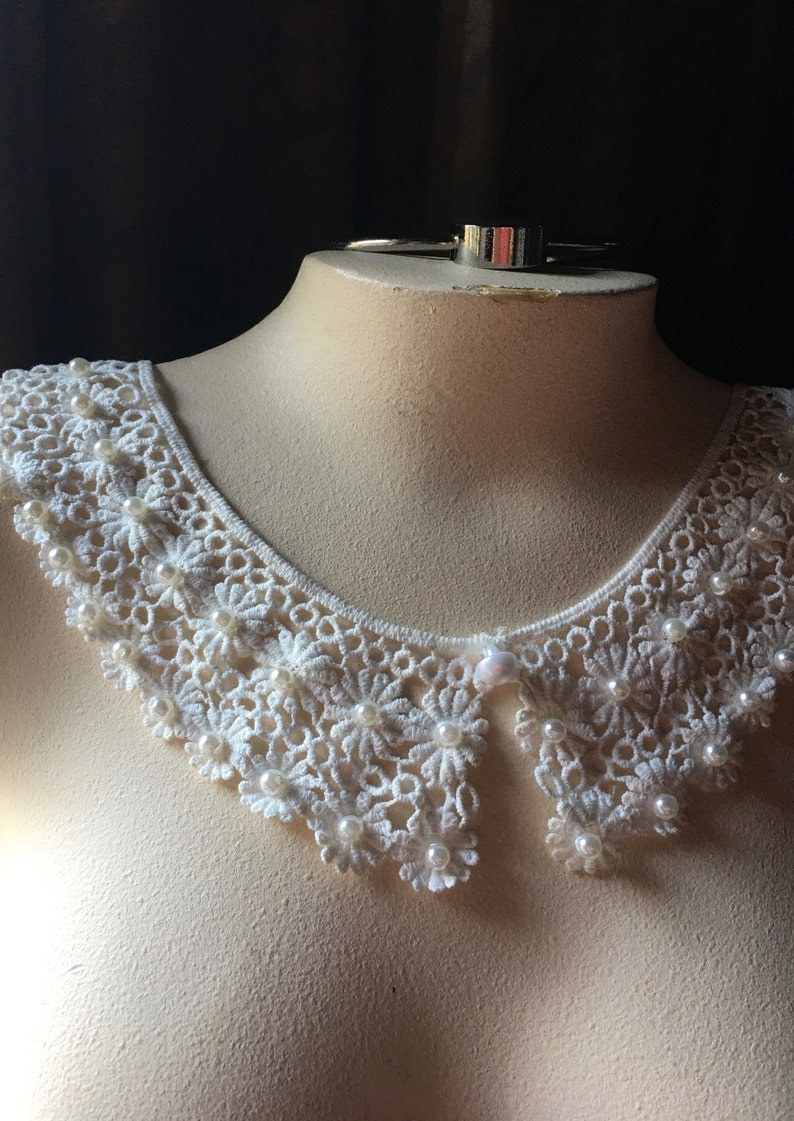 Puede incluir: Collar de encaje blanco con adornos de perlas. El collar est&aacute; en un busto de maniqu&iacute; blanco.