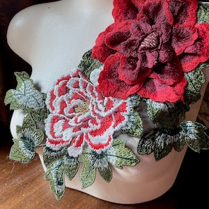 May include: A floral embroidered applique featuring two large flowers in shades of red and white, with green leaves. The applique is displayed on a mannequin, showcasing the intricate details of the embroidery.