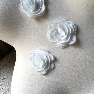 Puede incluir: Tres apliques de rosas blancas bordadas están dispuestos sobre una superficie color crema. Los delicados diseños florales presentan pétalos en capas, creando un efecto tridimensional. Estos apliques se pueden utilizar para manualidades o para embellecer prendas.