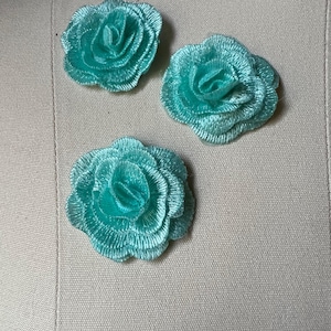 Puede incluir: Tres apliques de rosas bordadas en color turquesa. Los apliques están dispuestos sobre una superficie de tela beige. Cada rosa tiene múltiples capas de pétalos, creando un efecto tridimensional. El color es un turquesa vibrante y llamativo.