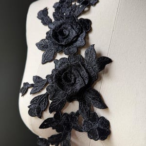 Pode incluir: Aplique de renda floral preta com desenhos de rosas e folhas. O aplique é exibido em um manequim, destacando o bordado detalhado. Ideal para embelezar roupas ou projetos de artesanato.