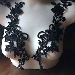 May include: Two black floral lace appliques are displayed on a white mannequin. The appliques feature intricate floral and leaf designs, with delicate details and a symmetrical arrangement. The lace is a deep black color, creating a striking contrast against the white background.