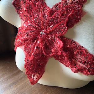 May include: A vibrant red sequined applique in a floral design. The applique is heavily embellished with sequins and features a central jewel. The design is displayed on a mannequin, showcasing its shape and texture.