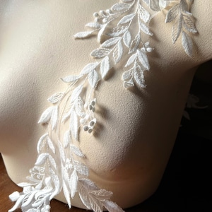 May include: Delicate white lace applique featuring a floral and leaf design. The applique is displayed on a mannequin, showcasing its intricate details and texture. Perfect for bridal wear or other craft projects.