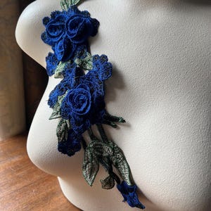 Puede incluir: Una aplicación floral azul y verde con tres rosas azules grandes y hojas verdes. La aplicación está sobre un fondo blanco.