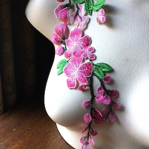 May include: A floral embroidered applique featuring pink flowers with green leaves and black stems. The applique is designed to cascade down, showcasing detailed stitching and a delicate aesthetic. Ideal for embellishing garments or crafts.