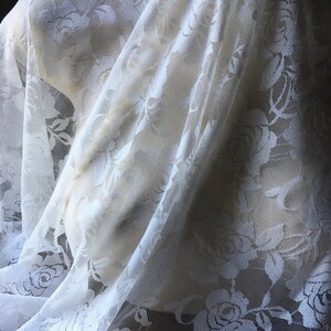 May include: White lace fabric with a floral rose pattern. The delicate material is draped over a mannequin, showcasing the intricate design and texture. The lace is semi-transparent, allowing light to filter through.