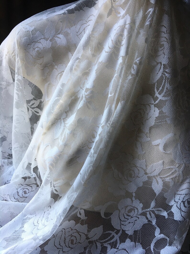 May include: White lace fabric with a floral rose pattern. The delicate material is draped over a mannequin, showcasing the intricate design and texture. The lace is semi-transparent, allowing light to filter through.