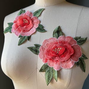 May include: Two embroidered floral patches featuring layered pink and red peony-style blossoms with dark green leaves. The petals exhibit a gradient of pink tones, while the centers are accented with small, metallic gold-colored thread details.