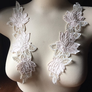 May include: Two pale pink floral lace appliques with intricate white detailing. The appliques are arranged vertically, featuring layered petals and delicate leaf accents. They are displayed on a mannequin, showcasing their potential use in garment design.