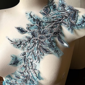 May include: A teal and navy blue floral applique with intricate bead and sequin detailing. The design features leaves and flowers on a sheer mesh backing. The applique is displayed on a white mannequin, showcasing its potential use in fashion or craft projects.