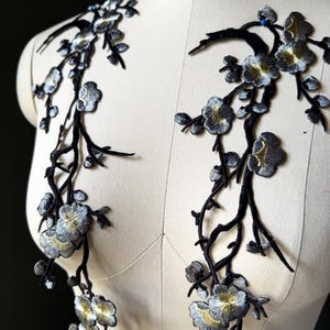May include: Two symmetrical floral embroidery appliques featuring black branches with silver and gold metallic thread petals. The detailed botanical design showcases blooming flowers and buds, crafted with intricate stitching to create a textured appearance.
