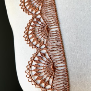 May include: Close-up of a decorative brown lace trim. The lace features a repeating pattern of circular and diamond-shaped designs. The trim is displayed against a white background, highlighting its intricate details. Ideal for sewing and craft projects.