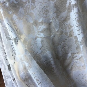 May include: Close-up of a white lace fabric with a floral rose pattern. The delicate material is draped, showing the intricate details of the design. The fabric is a light cream color, with a sheer, open weave.