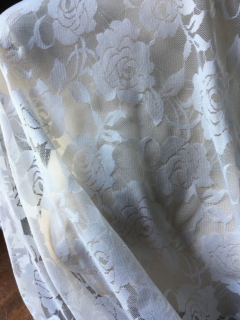 May include: Close-up of a white lace fabric with a floral rose pattern. The delicate material is draped, showing the intricate details of the design. The fabric is a light cream color, with a sheer, open weave.