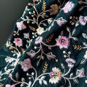 以下が含まれることがあります： 濃い緑色の生地に施された、複雑な花の刺繍のクローズアップ。刺繍は、ピンク、黄色、金色の花と銀色のアクセントが特徴です。生地は折りたたまれ、影を作り、デザインの質感と細部を際立たせています。