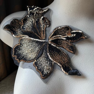 Puede incluir: Una aplicación floral negra y dorada con lentejuelas. La aplicación está sobre una tela negra transparente.