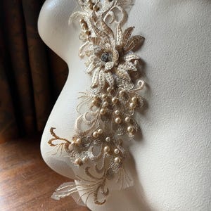 May include: Gold lace applique with pearl beads and a rhinestone embellishment. This delicate applique is perfect for adding a touch of elegance to wedding dresses, bridal accessories, or other special occasion garments.