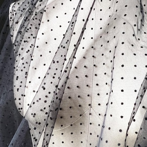 May include: Black tulle fabric with small, evenly spaced black polka dots. The sheer material is draped over a white dress form, creating soft folds and shadows. The fabric's texture and pattern are clearly visible.