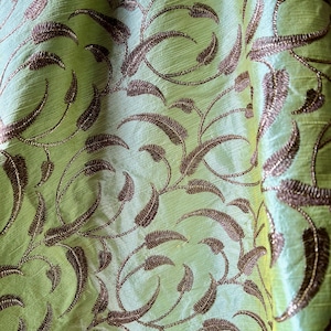 May include: Close-up of a light green fabric with a repeating pattern of brown embroidered leaves and vines. The fabric has a subtle sheen, and the embroidery adds texture and depth to the design. The overall effect is elegant and decorative.