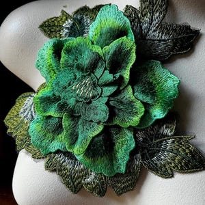May include: Green embroidered flower applique with a layered design. The flower has a green and white ombre effect. The applique is perfect for adding a touch of elegance to clothing, bags, or home decor projects.