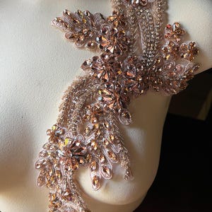 May include: A rose gold and clear crystal applique in a floral design. The applique features various sizes of crystals and beads, creating a sparkling effect. It is mounted on a sheer, light pink fabric, suitable for embellishing garments or accessories.