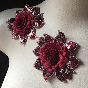 May include: Two burgundy red fabric flower appliques with sequins and a mesh backing. The flowers are in a rose shape and have a 3D effect.