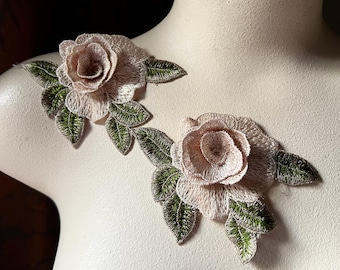 Puede incluir: Dos delicados apliques de tela de rosa de color rosa claro con hojas verdes. Las rosas están superpuestas y tienen un aspecto ligeramente texturizado.