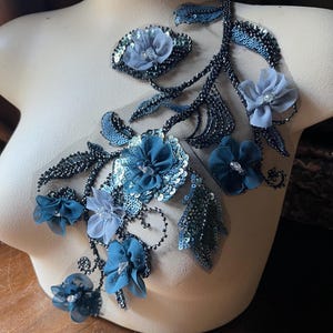 Könnte beinhalten: Eine blaue und silberne Blumenapplikation mit Pailletten und Perlen. Die Applikation zeigt verschiedene Blumenformen und -größen in einem filigranen, komplizierten Design.