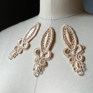 Może przedstawiać: Trzy beżowe aplikacje koronkowe z owalnym środkiem i ozdobnymi, wirującymi dolnymi detalami. Aplikacje są prezentowane na manekinie, prezentując ich delikatną fakturę i design. Aplikacje te nadają się do ozdabiania odzieży lub projektów rzemieślniczych.