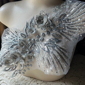 Puede incluir: Aplique floral plateado y blanco con lentejuelas y cuentas sobre una tela de malla transparente. El aplique presenta una flor grande con flores más pequeñas y hojas que la rodean.
