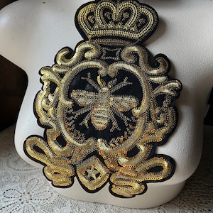 May include: A gold and black embroidered patch featuring a crown, ornate scrollwork, and a bee at its centre. The patch is displayed on a white mannequin.