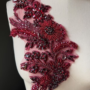May include: A burgundy beaded applique with a floral design. The applique features a variety of red beads, sequins, and crystals, arranged in a detailed pattern on a sheer mesh backing. The design includes a large central flower and several smaller floral elements.
