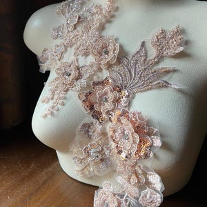 May include: A delicate floral lace applique with a rose gold color scheme. The applique features intricate floral designs, sequins, and beads. It is perfect for adding a touch of elegance to any garment.