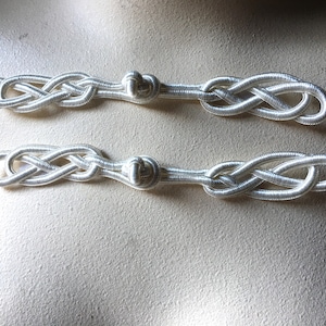 May include: Two silver-colored, decorative cord ornaments. Each ornament features a woven knot design on both ends, connected by a central knot. The cords are made of a shiny, reflective material.