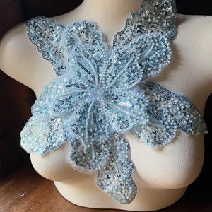 Pode incluir: Aplique floral azul claro com lantejoulas e contas, exibido em um busto de manequim. O design floral tem várias camadas de pétalas e um aglomerado central de contas.