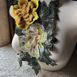 May include: A pair of yellow and white embroidered flowers with green leaves. The flowers are attached to a white fabric background.