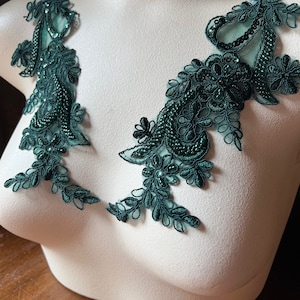 Puede incluir: Dos apliques de encaje verde oscuro con diseños florales y con cuentas. Los apliques se muestran en un maniquí, mostrando sus intrincados detalles y su potencial de embellecimiento. El encaje presenta un delicado patrón calado.