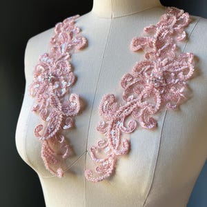 May include: Two pink lace appliques with floral and swirl designs. The appliques are embellished with sequins and small pearls. The lace is displayed on a mannequin, showcasing the delicate details and texture of the fabric.
