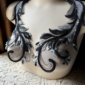 Op de afbeelding: Twee zwarte en zilveren bloemvormige kanten applicaties met pailletten, getoond op een witte mannequin.