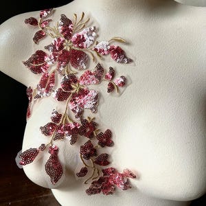 Op de afbeelding: Een bloemenapplicatie met bordeauxrode en zilveren pailletten en gouddraad details. Het ontwerp toont een delicaat, gelaagd bloemmotief, perfect voor het verfraaien van kleding of accessoires. De pailletten vangen het licht en voegen glans toe.