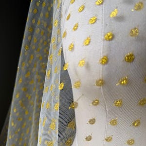 Könnte beinhalten: Ein transparenter weißer Tüllstoff mit einem sich wiederholenden Muster aus kleinen, runden goldenen Glitzerpunkten. Das leichte, halbtransparente Material ist über eine Schneiderpuppe drapiert und zeigt die Textur sowie die goldenen Verzierungen.