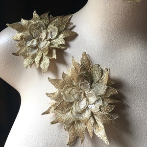 Pode incluir: Dois apliques de flores de tecido dourado com lantejoulas. As flores estão sobre um fundo castanho claro.