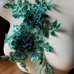 Peut inclure: Applique florale vert émeraude avec des paillettes et des détails brodés. L'applique présente de multiples grappes de fleurs et des motifs de feuilles. Les paillettes ajoutent un effet scintillant au tissu. L'applique est présentée sur un mannequin.